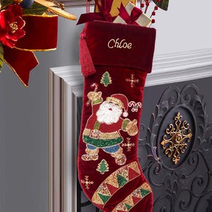 Puede incluir: Un calcetín navideño de terciopelo rojo intenso con una figura de Papá Noel hecha de cuentas, copos de nieve bordados y un patrón decorativo de cuentas. El nombre Chloe está bordado en hilo dorado en el puño, que incluye una borla roja.
