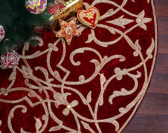 May include: A round, deep red Christmas tree skirt with gold embroidered floral and vine patterns. The skirt is made of a soft velvet material and is decorated with gold beads. Ornaments and a gift box are visible.