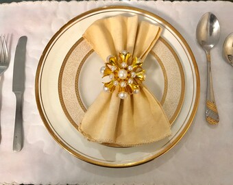 May include: A white dinner plate with a gold rim and a delicate floral pattern. A beige napkin is folded and placed on the plate, secured with a gold and pearl napkin ring. Two silver spoons and a silver knife are placed on a white tablecloth.