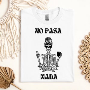 May include: White t-shirt with the text "NO PASA NADA" above a skeleton graphic wearing sunglasses and holding a coffee mug. The skeleton is giving a peace sign and is surrounded by flowers. The text "NADA" is below the graphic.