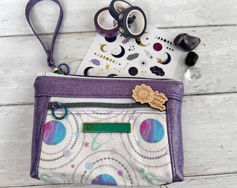 May include: Purple zippered pouch with a white and purple galaxy print. The pouch has a zippered pocket on the front and a small wooden tag that says "hidden gems & delights".