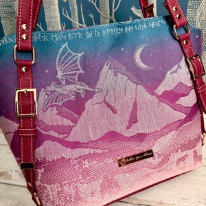 May include: A tote bag with a fantasy landscape design. The bag features a dragon flying over mountains under a moonlit sky, with a color gradient from blue to pink. It has magenta straps and gold-colored buckles.