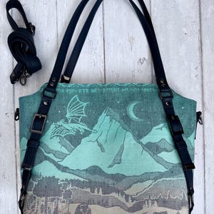 May include: A teal tote bag with a mountain and dragon design. The bag has black straps and a detachable shoulder strap. The design features a dragon, mountains, a moon, and forest animals. The bag is made of a woven fabric.