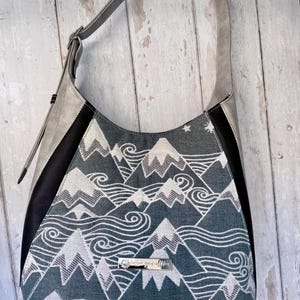 May include: A gray and black shoulder bag with a mountain and wave pattern. The bag has a gray strap and a silver accent. The mountain design is white and gray on a blue-gray background. The bag has a silver nameplate.
