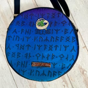 May include: A round blue and purple crossbody bag with a black strap. The bag is covered in black runes and has a gold ring with a green leaf design attached to it. The bag has a gold label that says "little girl's oddities".