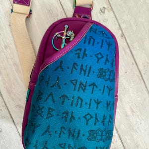 May include: A purple and blue crossbody bag with a  leather strap and a  metal pin with a sword and flower design. The bag is covered in a pattern of runes.