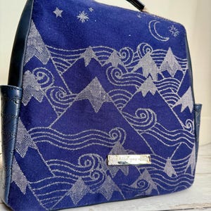 May include: A navy blue backpack with a silver mountain and wave design. The backpack has a silver metal tag with the text "little  goes  a  long  way".