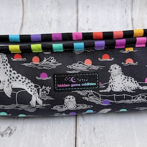 May include: A black pencil case with a colorful striped zipper and handle. The case features a repeating pattern of seals, waves, and sunsets. The text "hidden gems oddities" is on a small patch.