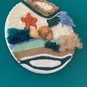 Pode incluir: Uma parede redonda feita de fio e tecido em tons de branco, marrom, azul, verde e laranja. A parede apresenta uma estrela do mar, conchas e uma variedade de texturas.