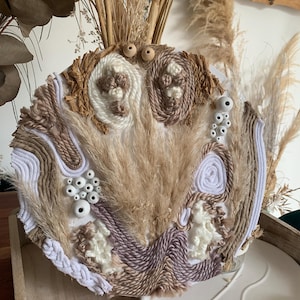 May include: A circular wall hanging made of woven natural fibers in shades of brown, white, and beige. The hanging features a variety of textures, including fluffy fibers, rope, and small white beads.