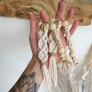 Puede incluir: Tres llaveros hechos a mano. Los dos primeros son de macramé con detalles dorados y borlas. El tercer llavero tiene cuentas de madera con la palabra "ROMANE". Todos los llaveros se sostienen en una mano.