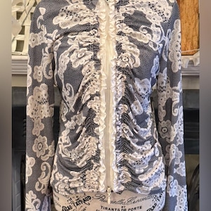 May include: A black and white patterned jacket with a white lace overlay. The jacket has a high collar, long sleeves, and a full-length zipper. The front features a ruffled white panel. The jacket is displayed on a mannequin.