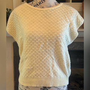 May include: A short-sleeved, light yellow knit top with a textured pattern. The top has a crew neck and a ribbed hem. The fabric appears to be a soft, breathable material, suitable for warmer weather. The overall style is casual and comfortable.