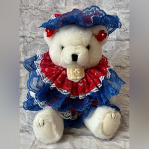 May include: A white teddy bear wearing a patriotic outfit. The outfit includes a blue lace hat, a red and blue lace collar, and a matching dress. The bear has a black nose and small red bows on its ears.