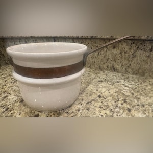 May include: A white ceramic saucepan with a copper-colored band around the middle and a long metal handle. The saucepan has a rounded shape and a wide opening. The handle extends from the side of the pan.