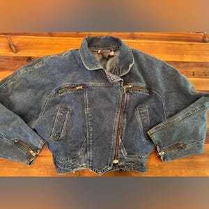 May include: A medium-wash blue denim jacket featuring an asymmetrical front zipper closure, a wide collar, and two horizontal zippered chest pockets. The jacket includes two vertical side pockets and zippers at the ends of the long sleeves.