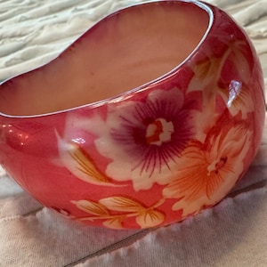 May include: A decorative, oval-shaped bowl with a glossy, pink exterior. The bowl is adorned with a floral pattern in shades of pink, white, and orange. The interior of the bowl is a soft peach color.