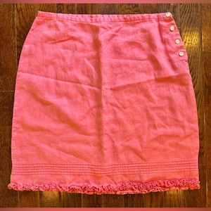 May include: A coral-colored skirt with a straight cut and a ruffled hem. The skirt features three white buttons on the side and horizontal stitching details near the hem. The fabric appears to be linen or a similar lightweight material.