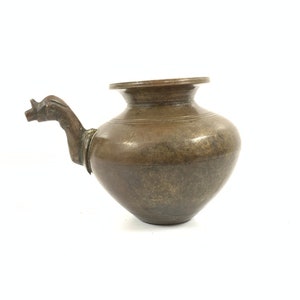 Op de afbeelding: Een kleine, antieke messing waterkan met een tuit in de vorm van een dierenkop. De kan heeft een rond lichaam en een brede, platte basis.