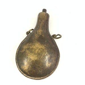 May include: A brass powder flask with a pear-shaped body and a small spout at the top. The flask has two metal loops on the side for attaching a strap.