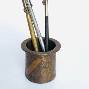 May include: A brass pen holder with an intricate design, featuring three pens: a silver and gold pen, a silver pen, and a black pen.