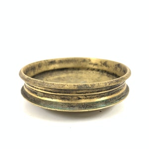 May include: A small, round, brass bowl with a slightly raised rim. The bowl has a worn, aged appearance with a greenish patina.