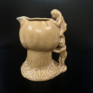 Op de afbeelding: Een keramische kan in de vorm van een paddenstoel met twee figuren die erop klimmen. De kan is lichtbruin van kleur en heeft een getextureerd oppervlak.