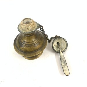 Puede incluir: Lámpara de aceite antigua de latón con una cadena y un portamechas separado. La lámpara tiene un cuerpo redondo con un diseño decorativo y un pequeño pico. El portamechas también está hecho de latón y tiene una forma redonda.