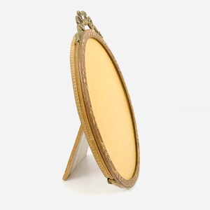 May include: An antique oval picture frame with a decorative brass finish. The frame has an ornate top design and a stand on the back. The frame is empty, with a yellow backing visible inside. The frame is angled slightly to the side.