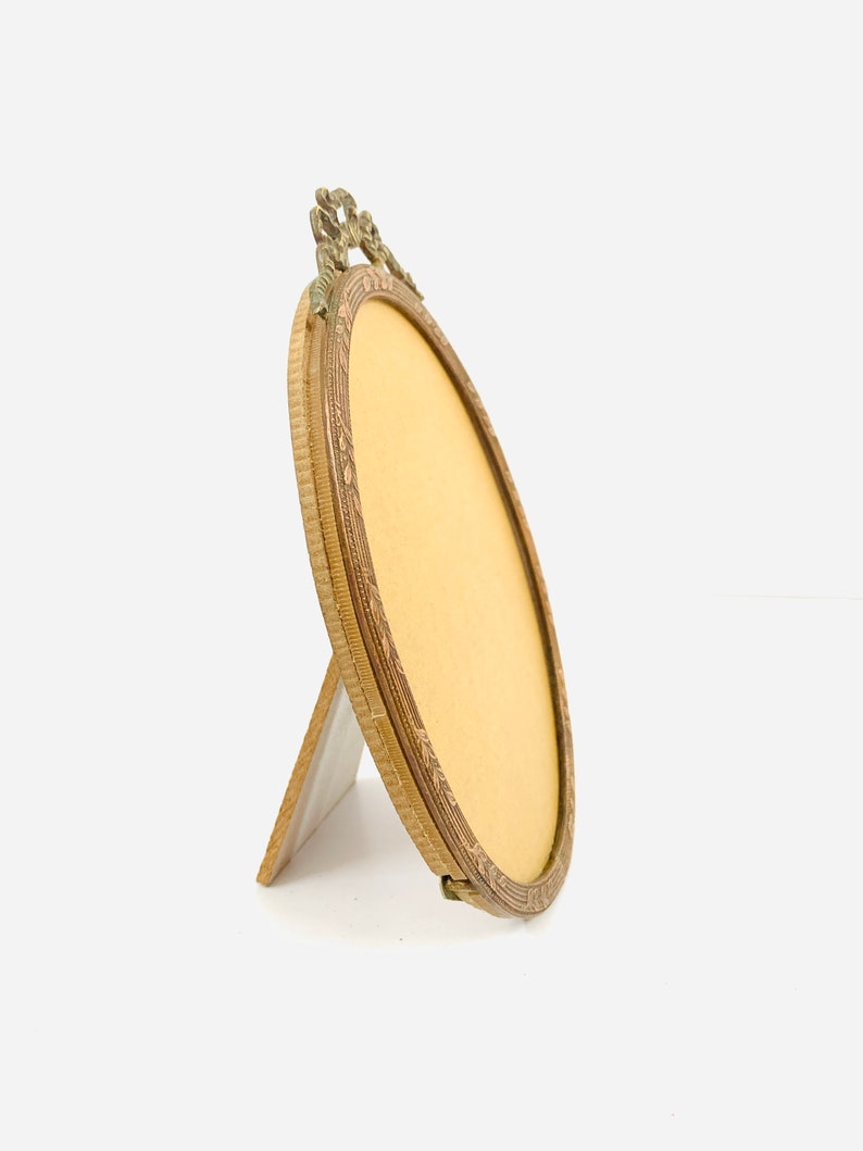 May include: An antique oval picture frame with a decorative brass finish. The frame has an ornate top design and a stand on the back. The frame is empty, with a yellow backing visible inside. The frame is angled slightly to the side.