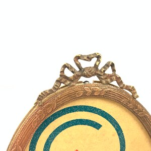 May include: Oval-shaped antique picture frame with a decorative bow at the top. The frame has a gold-colored finish with a detailed border. The background of the picture is tan with blue and red accents.