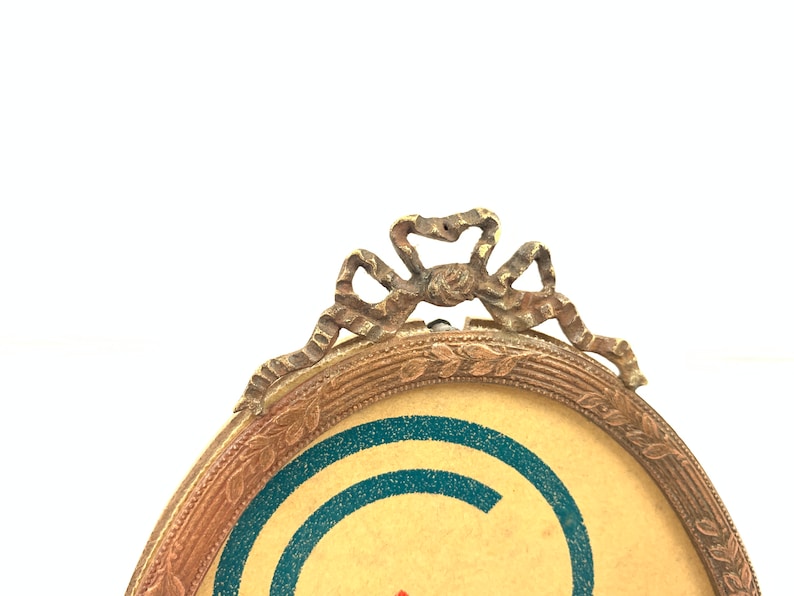 May include: Oval-shaped antique picture frame with a decorative bow at the top. The frame has a gold-colored finish with a detailed border. The background of the picture is tan with blue and red accents.