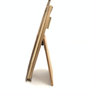 May include: A small, rectangular wooden picture frame with a decorative top. The frame is supported by a wooden easel back. The frame is a light brown color with a gold-colored metal trim.