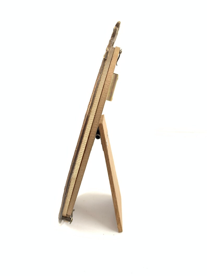 May include: A small, rectangular wooden picture frame with a decorative top. The frame is supported by a wooden easel back. The frame is a light brown color with a gold-colored metal trim.