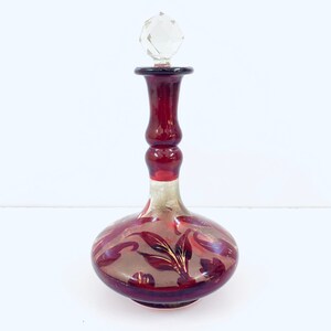 Op de afbeelding: Een rode glazen fles met een doorzichtige glazen stop. De fles heeft een gouden en wit bloemenmotief.