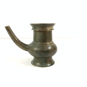 Könnte beinhalten: Ein dunkelgrüner Metallkrug mit einem Ausguss und einem runden Körper. Der Krug hat einen dekorativen Rand und einen leicht ausgestellten Boden.