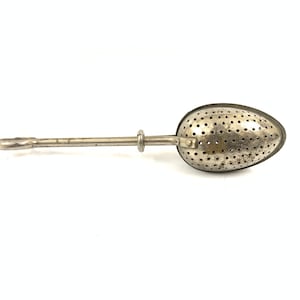 Op de afbeelding: Een zilveren thee-ei met een lange steel en een geperforeerde ovale mand.