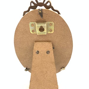 May include: Back view of an oval-shaped picture frame with a decorative bow at the top. The frame has a wooden backing and a metal stand. The frame is designed to stand upright on a flat surface.