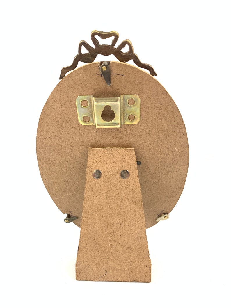 May include: Back view of an oval-shaped picture frame with a decorative bow at the top. The frame has a wooden backing and a metal stand. The frame is designed to stand upright on a flat surface.