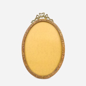 May include: Oval-shaped antique picture frame with a bronze finish. The frame features a decorative ribbon and leaf design at the top and around the border. The interior is a light tan color, ready for a photo.