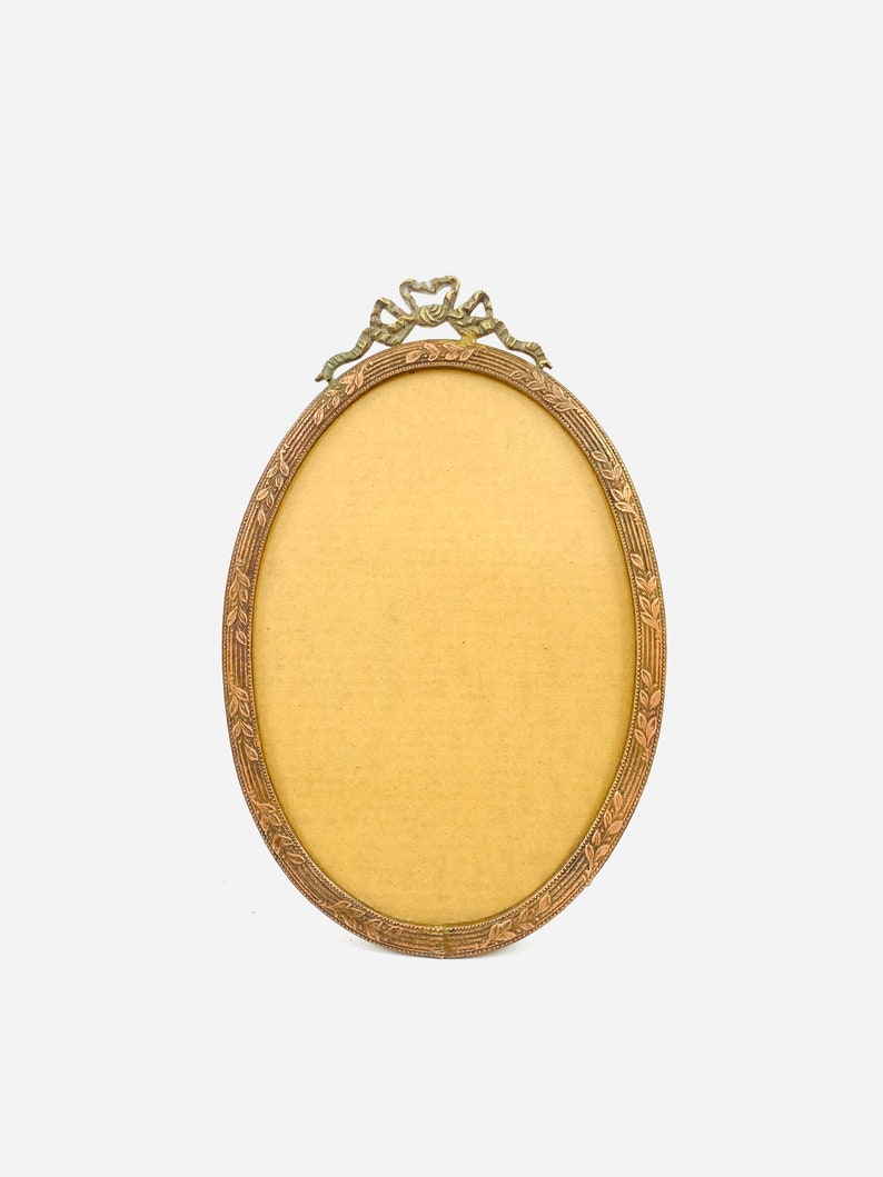 May include: Oval-shaped antique picture frame with a bronze finish. The frame features a decorative ribbon and leaf design at the top and around the border. The interior is a light tan color, ready for a photo.