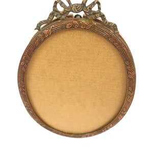May include: Oval-shaped antique picture frame with a bronze finish. The frame features a decorative bow at the top and a detailed leaf pattern around the inner edge. The frame's interior is a light tan color.