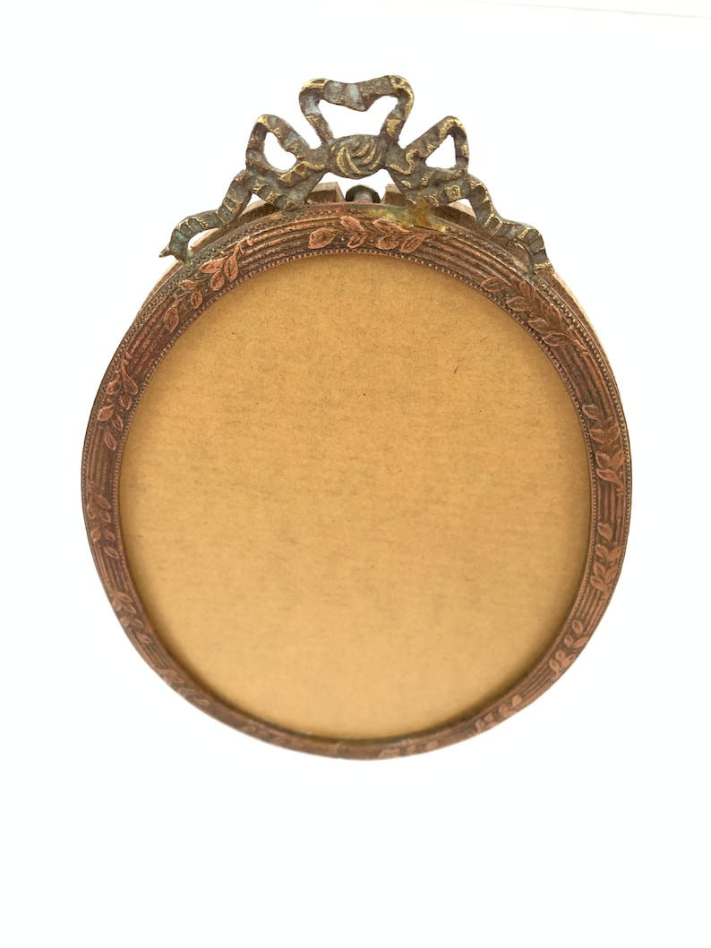 May include: Oval-shaped antique picture frame with a bronze finish. The frame features a decorative bow at the top and a detailed leaf pattern around the inner edge. The frame's interior is a light tan color.
