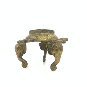 Może przedstawiać: Dekoracyjny stojak wykonany z pozłacanego metalu, z okrągłym blatem i trzema nogami. Każda noga ma kształt głowy słonia.