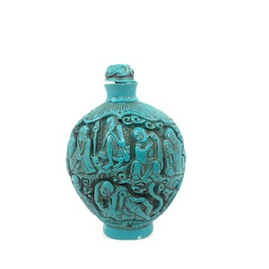 May include: A teal blue ceramic bottle with intricate carvings of figures and designs. The bottle has a round body and a small, round lid.