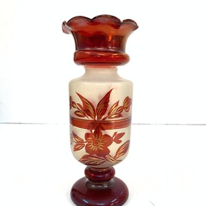 Puede incluir: Un pequeño jarrón de vidrio esmerilado con un diseño floral rojo y dorado. El jarrón tiene un borde festoneado y una base acampanada.