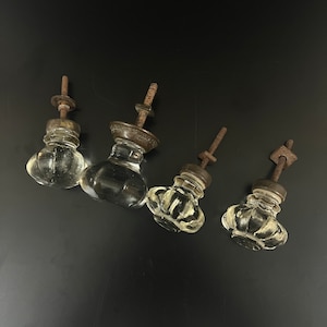 Puede incluir: Cuatro pomos de armario de vidrio antiguo con tallos de metal. Los pomos son de vidrio transparente con una superficie texturizada. Los tallos son de metal oxidado.