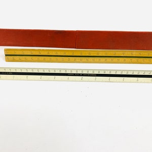 Könnte beinhalten: Zwei Lineale, eines aus Holz und eines in Schwarzweiß, mit einer roten Stoffhülle. Das Holzlineal ist in Zoll und das Schwarzweißlineal in Zentimetern gekennzeichnet.