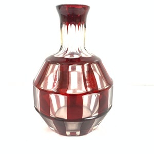 May include: A red glass decanter with a clear glass neck and a geometric pattern. The decanter has a round body and a wide base.