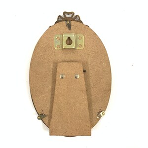 May include: Back of an oval-shaped picture frame with a decorative bronze-colored top. The frame has a cardboard backing with a metal hanger and a cardboard stand attached with small metal hinges and rivets.